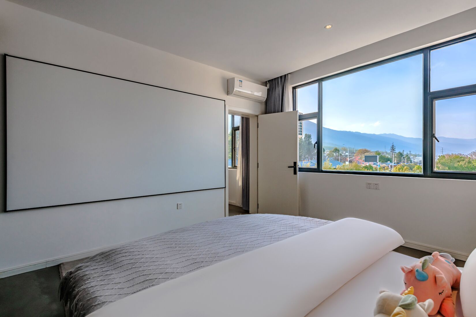 A modern hotel room with a large bed, a flat-screen TV, a large window with a city and mountains view, and a door leading to another room.
