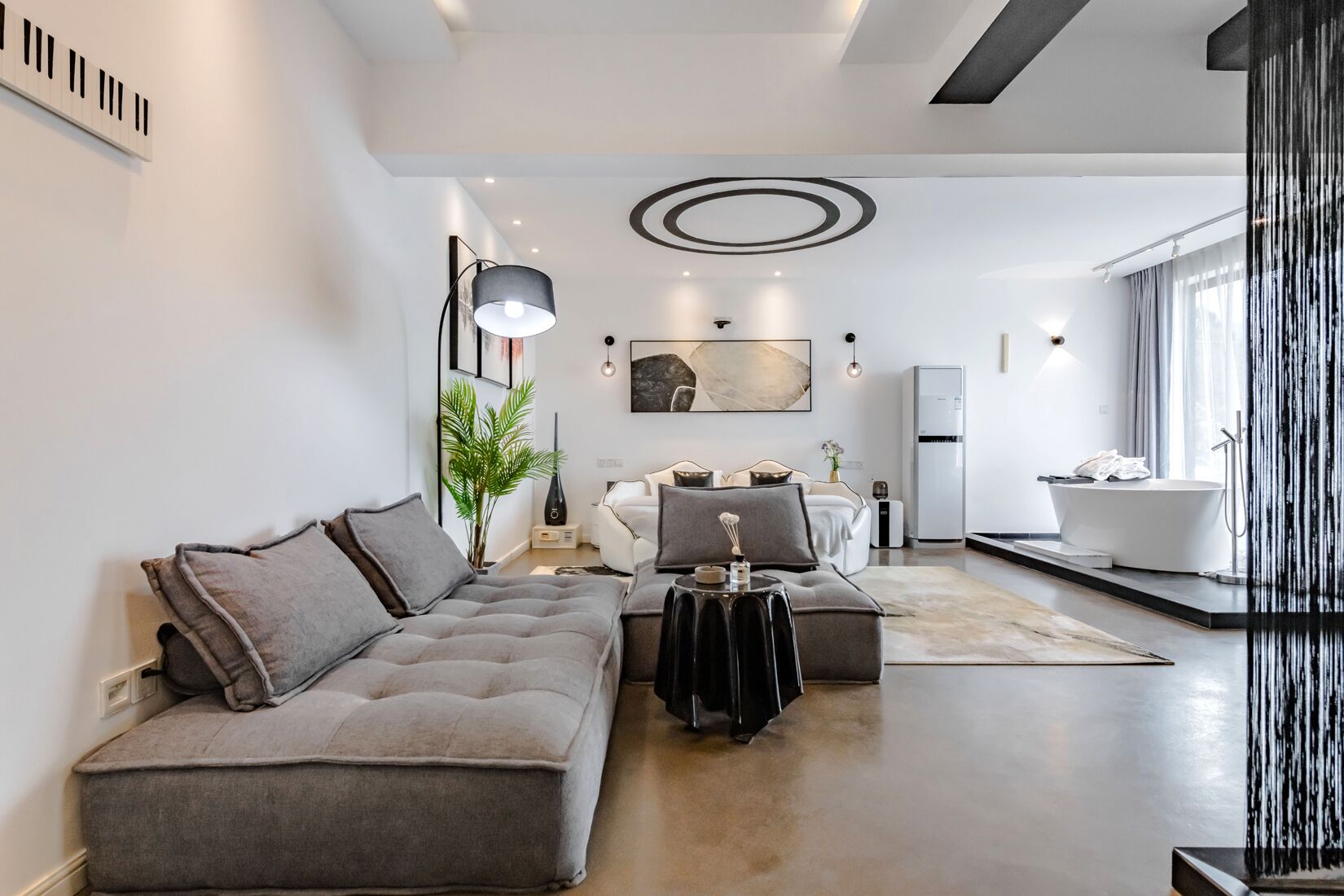 Ein modernes Wohnzimmer mit einem grauen Sofa, einem Couchtisch und einem Essbereich mit einem weißen Tisch und Stühlen. Es gibt eine Küchenzeile mit einem Kühlschrank und einer weißen Badewanne.