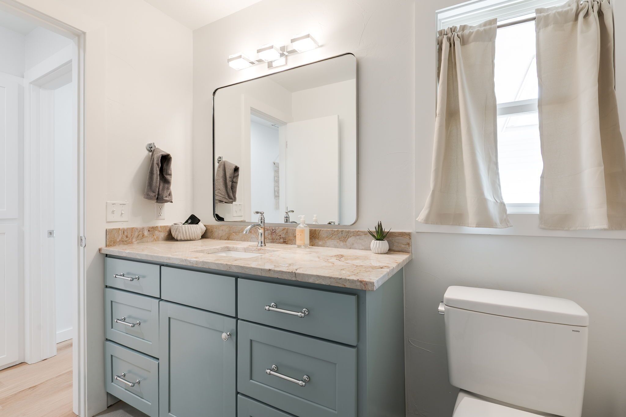 A bathroom with a marble countertop, a sink, a mirror, a toilet, and a window with curtains.