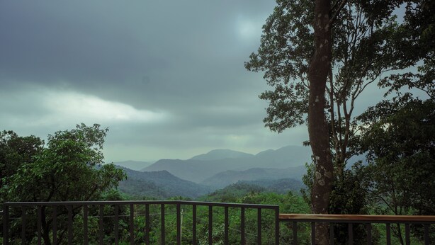 A view from a balcony with a railing, overlooking a forested landscape and distant mountains under a cloudy sky.