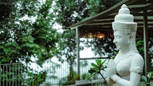 A white statue of a figure with hands in a prayer position, set against a backdrop of lush greenery and a building with a covered porch.