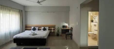 A hotel room with a bed, a desk, a chair, a ceiling fan, and a view of the bathroom.