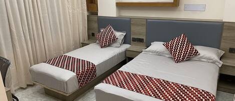 Two single beds with patterned bedspreads, a wooden headboard, a white curtain, and a light switch on the wall.