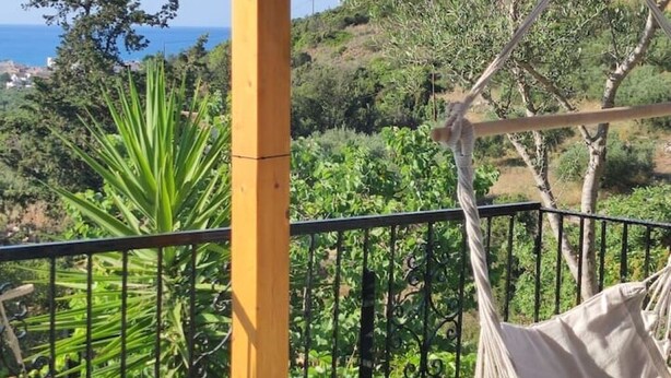 A balcony with a hammock, a yellow pole, and a view of a vineyard and the sea.
