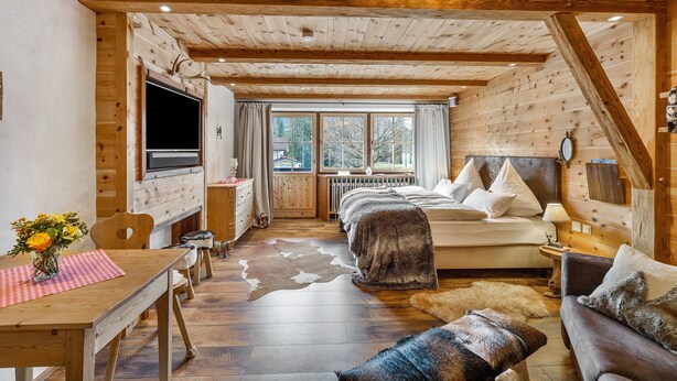 A log cabin interior with a wooden ceiling, a bed, a TV, a dining table with a vase of flowers, and a window with curtains.