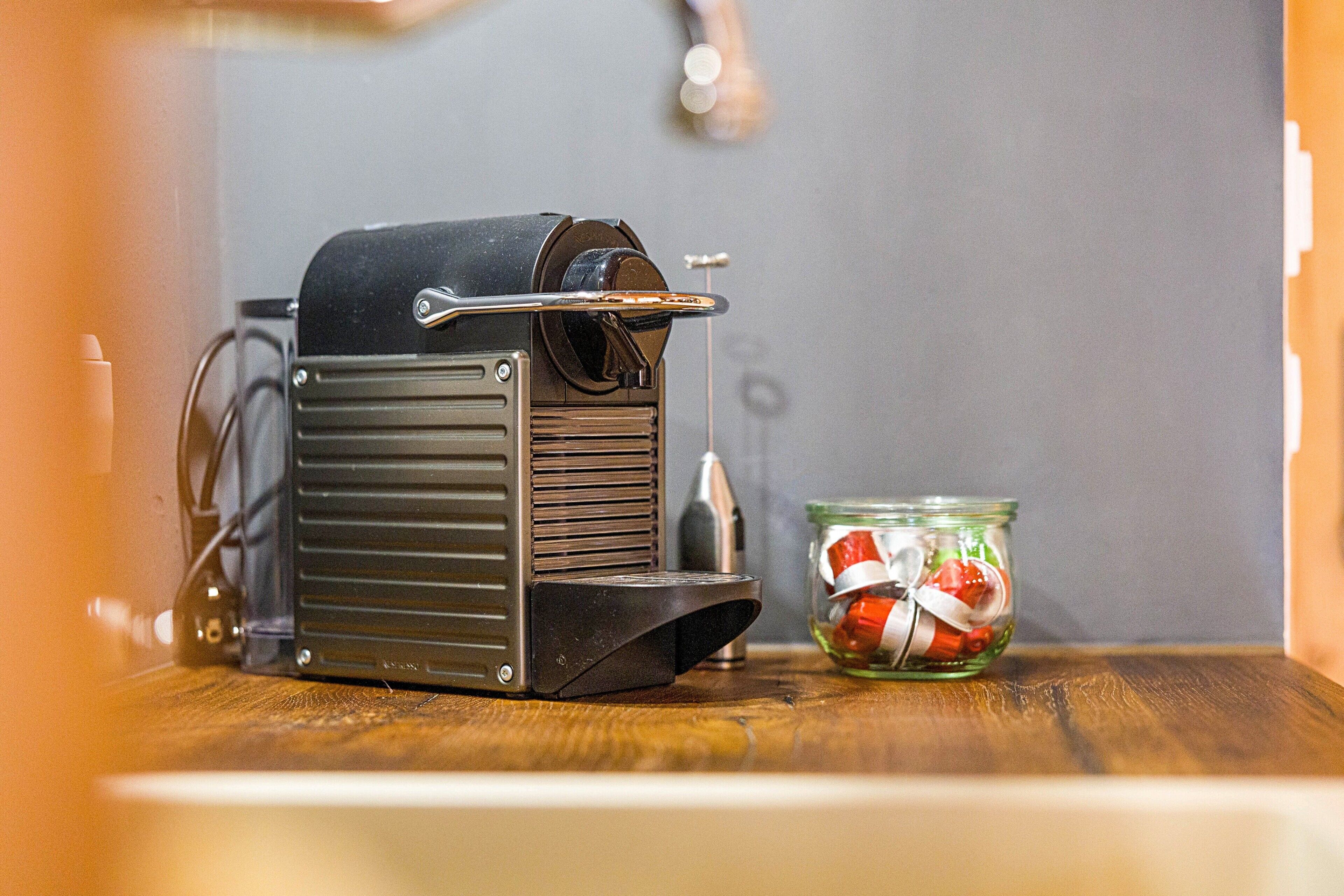 A coffee machine on a wooden surface with a glass jar containing red and white candies in the background.