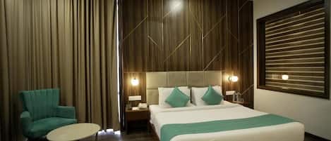 Een hotelkamer met een groot bed, een groene fauteuil, een klein rond tafeltje en een raam met jaloezieën.