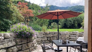 Une terrasse extérieure avec un sol en pierre, une table avec des chaises, un grand parasol et une vue sur un paysage montagneux.