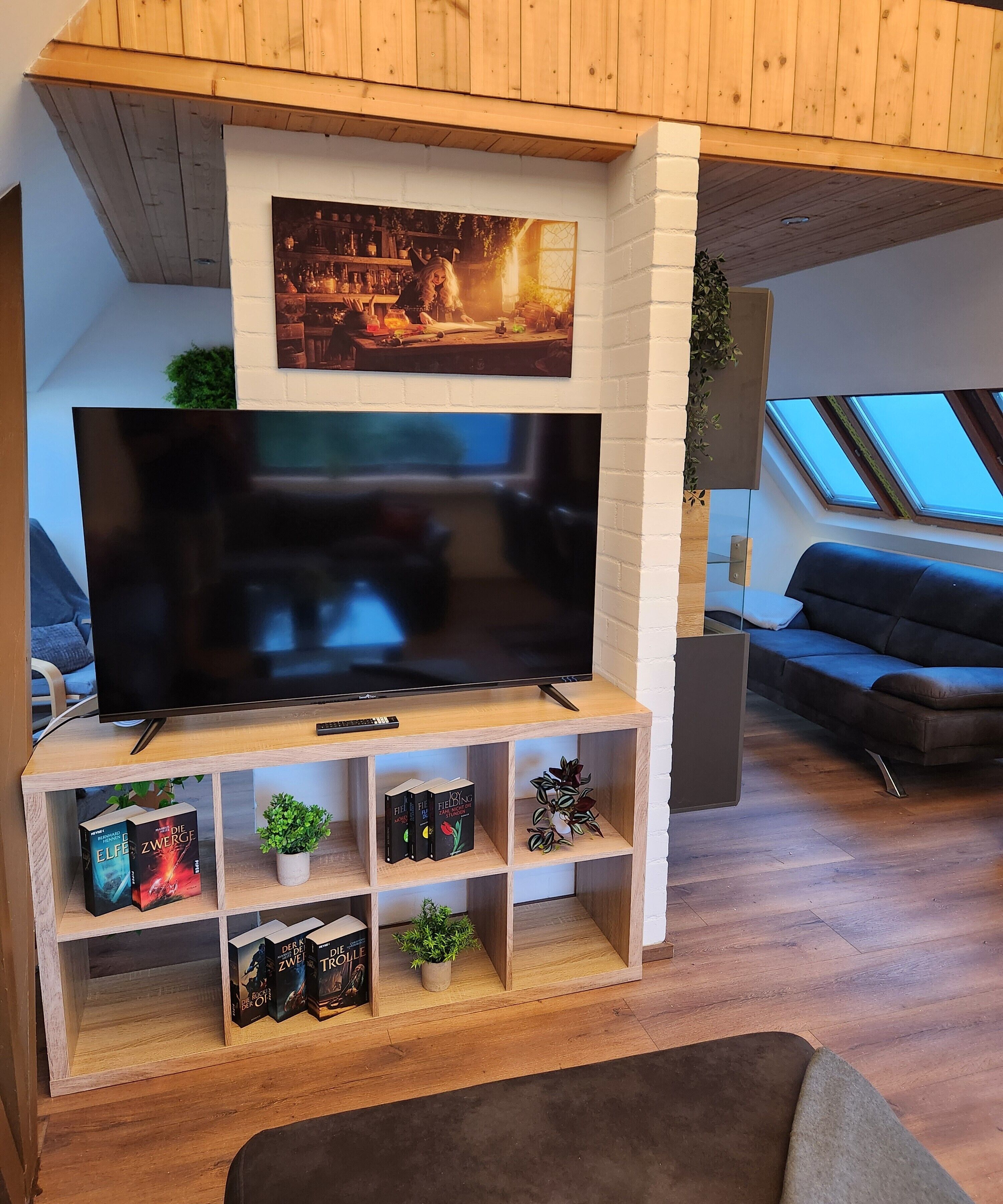 Una sala de estar con una televisión de pantalla grande sobre un mueble de madera, una estantería con libros, una planta pequeña y un sofá.