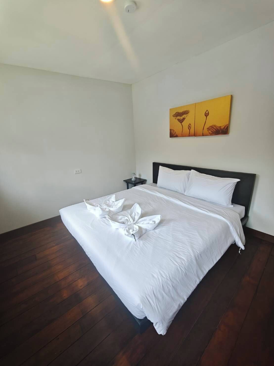 A hotel room with a bed, a nightstand, a painting on the wall, and wooden flooring.