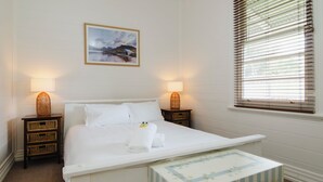 A neatly arranged bedroom with a bed, two bedside tables with lamps, a small bedside table with a basket, a framed picture on the wall, and a window with blinds.