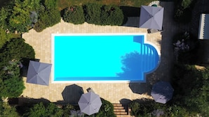 An aerial view of a rectangular swimming pool surrounded by a paved deck, with four umbrellas and a small staircase leading to a covered area.