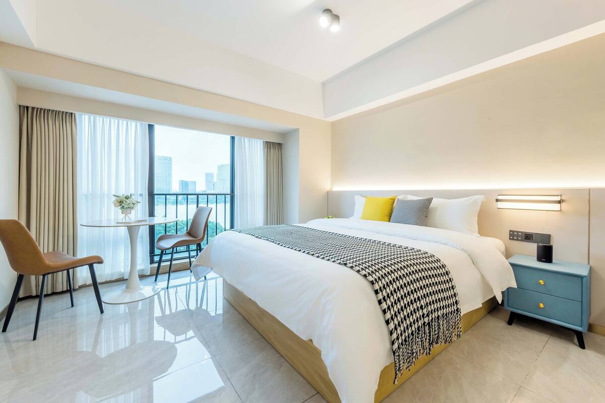 Une chambre d’hôtel moderne, dotée d’un grand lit, d’une table de chevet avec une lampe, d’une chaise et d’un balcon avec des rideaux.
