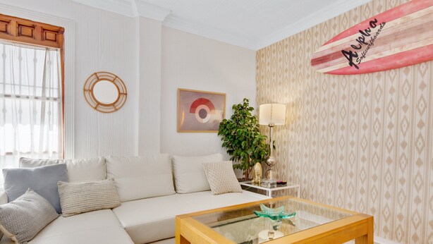 Ein Wohnzimmer mit beiger Couch, einem Glastisch, einer Surfbrettr-Dekoration, einem Spiegel und einer Wand mit Muster.