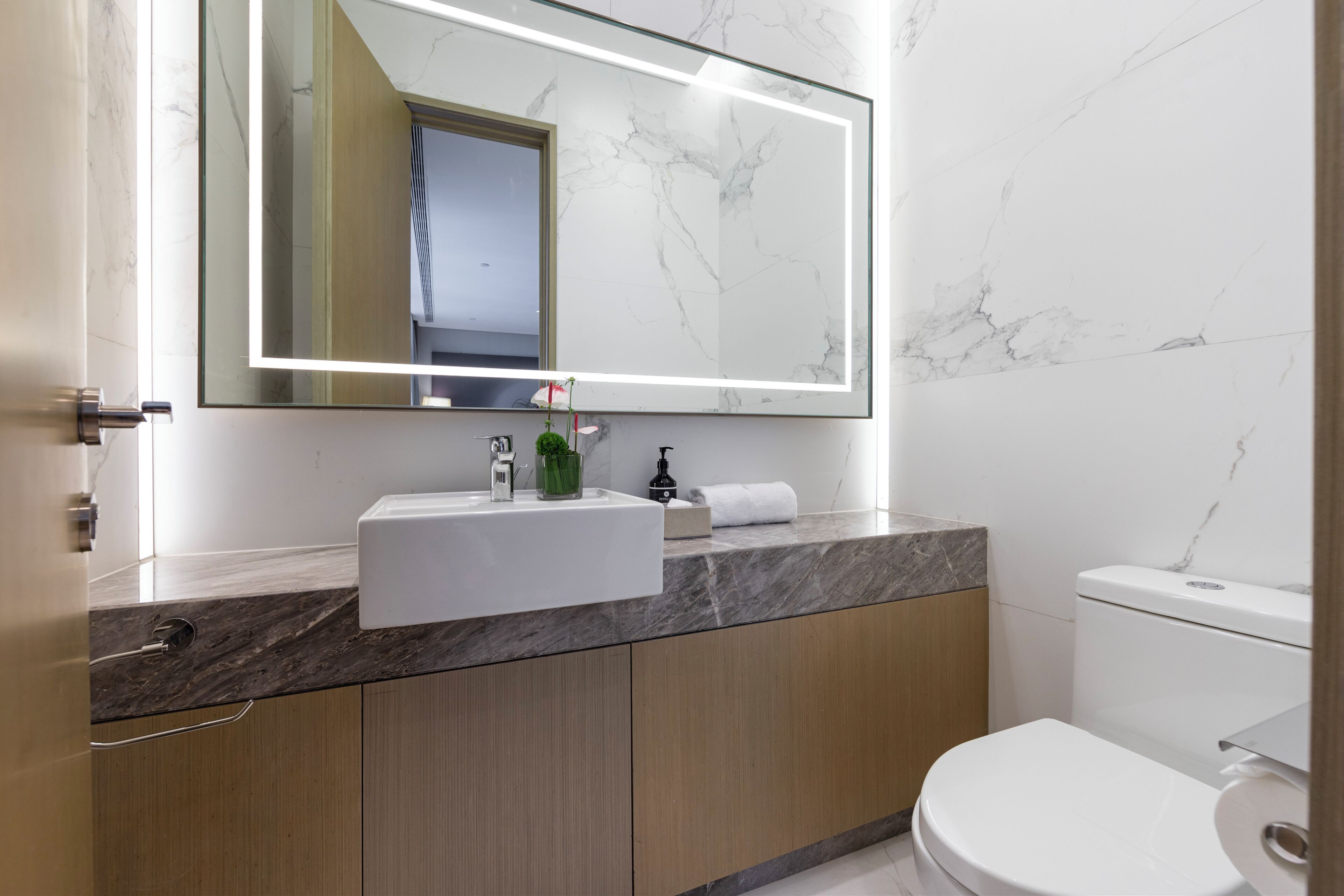 Un baño moderno con lavamanos de mármol, armarios de madera, un inodoro y un espejo grande con iluminación debajo del mueble.