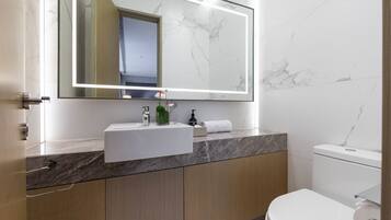Un baño moderno con lavamanos de mármol, armarios de madera, un inodoro y un espejo grande con iluminación debajo del mueble.