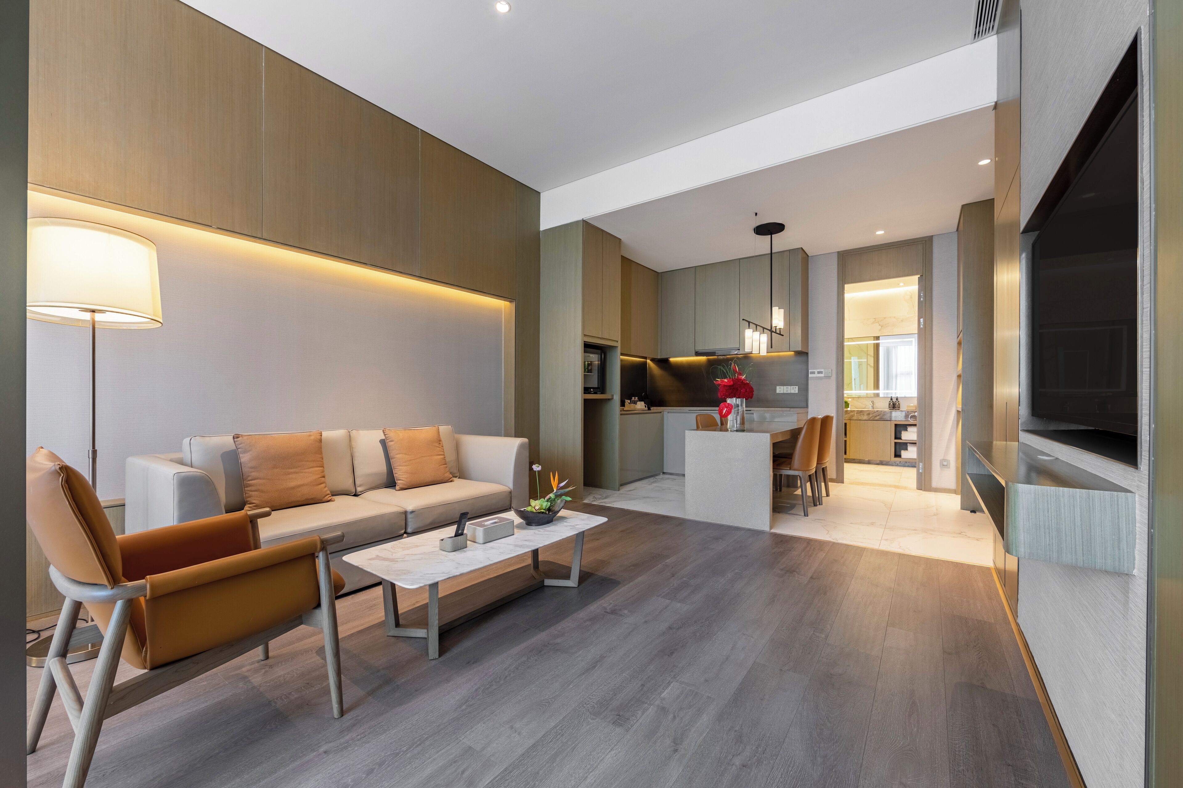 A modern living room with a sofa, coffee table, and a lamp. There is a kitchen area in the background with a dining table and chairs.