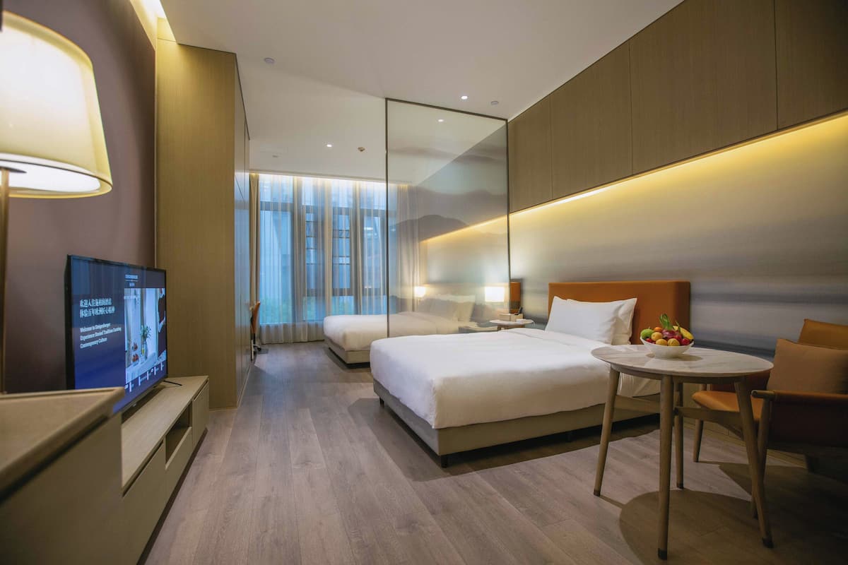 Habitación de hotel con cama, televisor, mesita de noche, silla y una pequeña mesa con fruta.