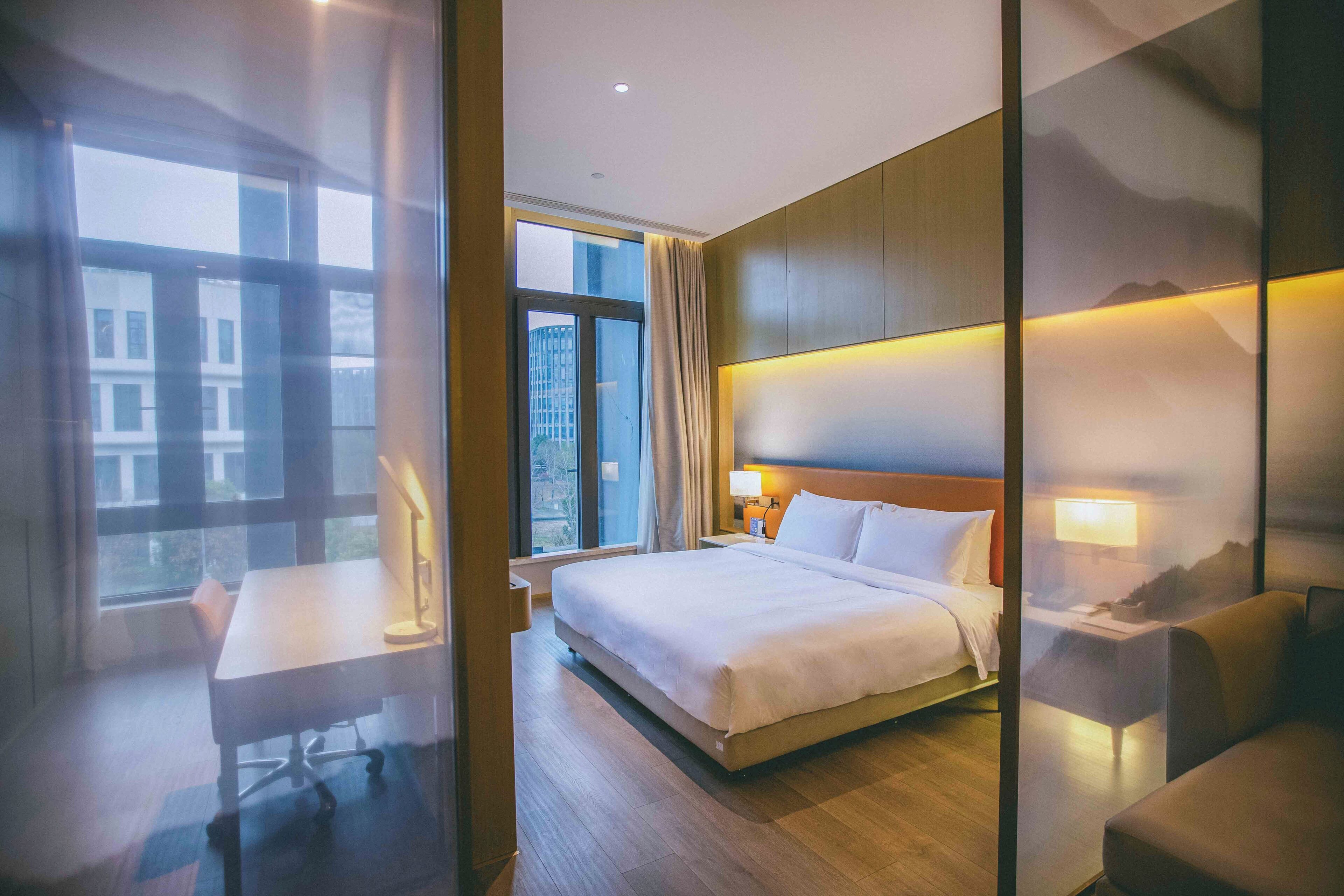 A hotel room with a large bed, a desk, a chair, a nightstand, and a view of the city through the window.