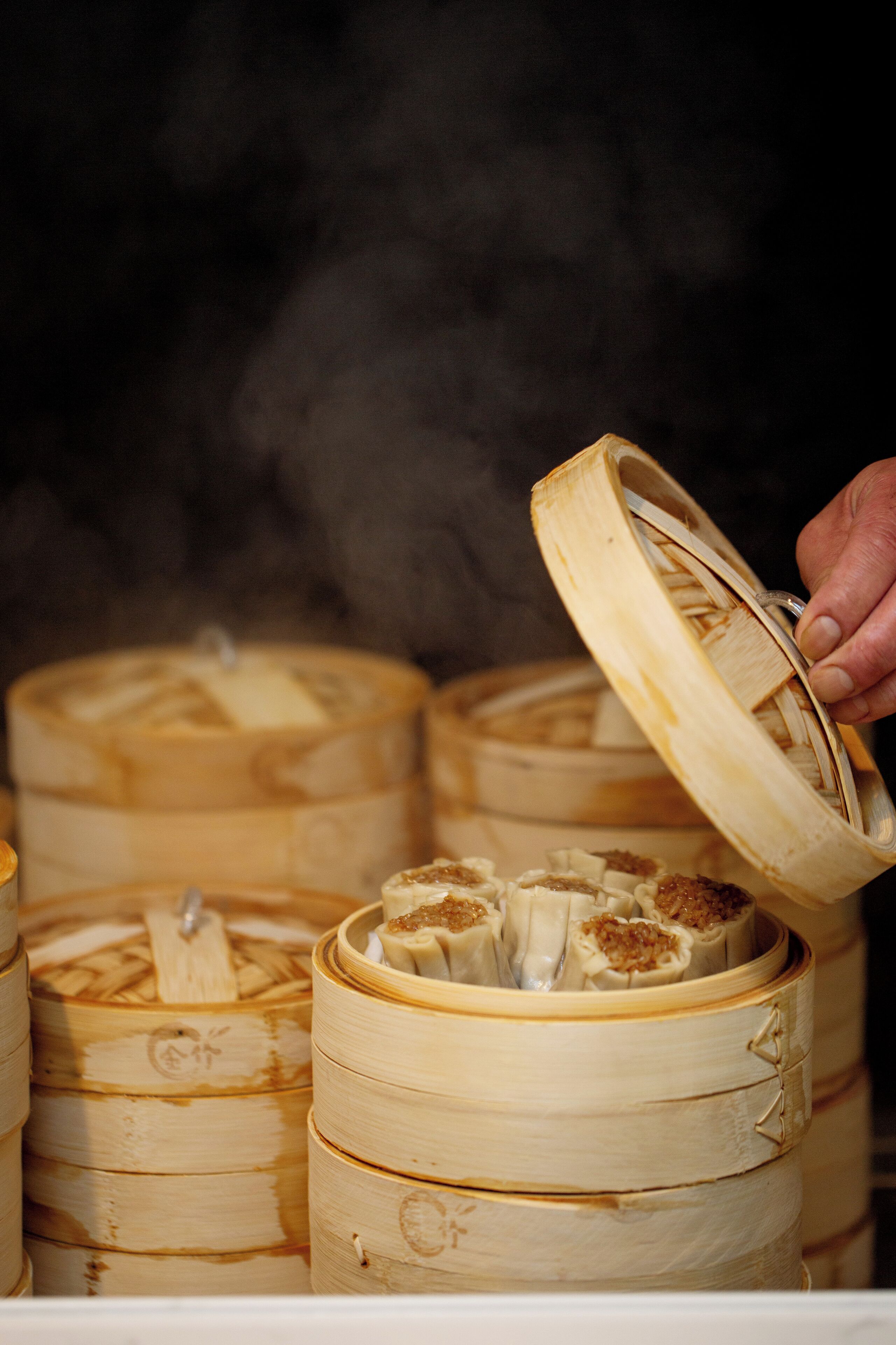 A hand lifting the lid of a bamboo steamer containing dumplings.