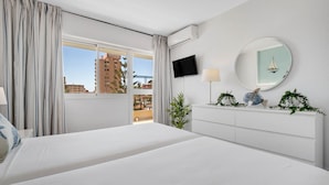 A hotel room with a large bed, a TV mounted on the wall, a round mirror, and a window with curtains.