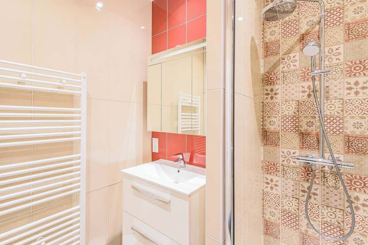 Une salle de bain avec un lavabo blanc, un miroir, une douche équipée d’un pommeau de douche à main et des carreaux décorés.