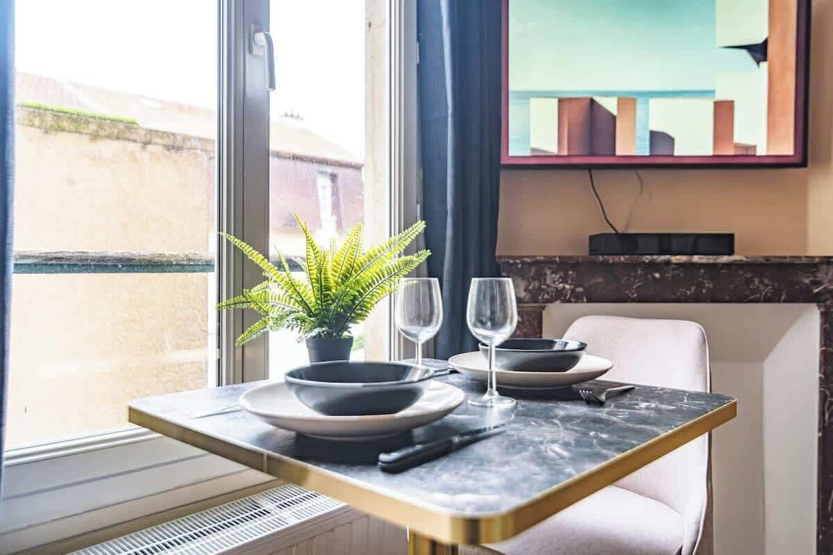 A dining area with a table set for two, a potted plant, and a view of the outside.