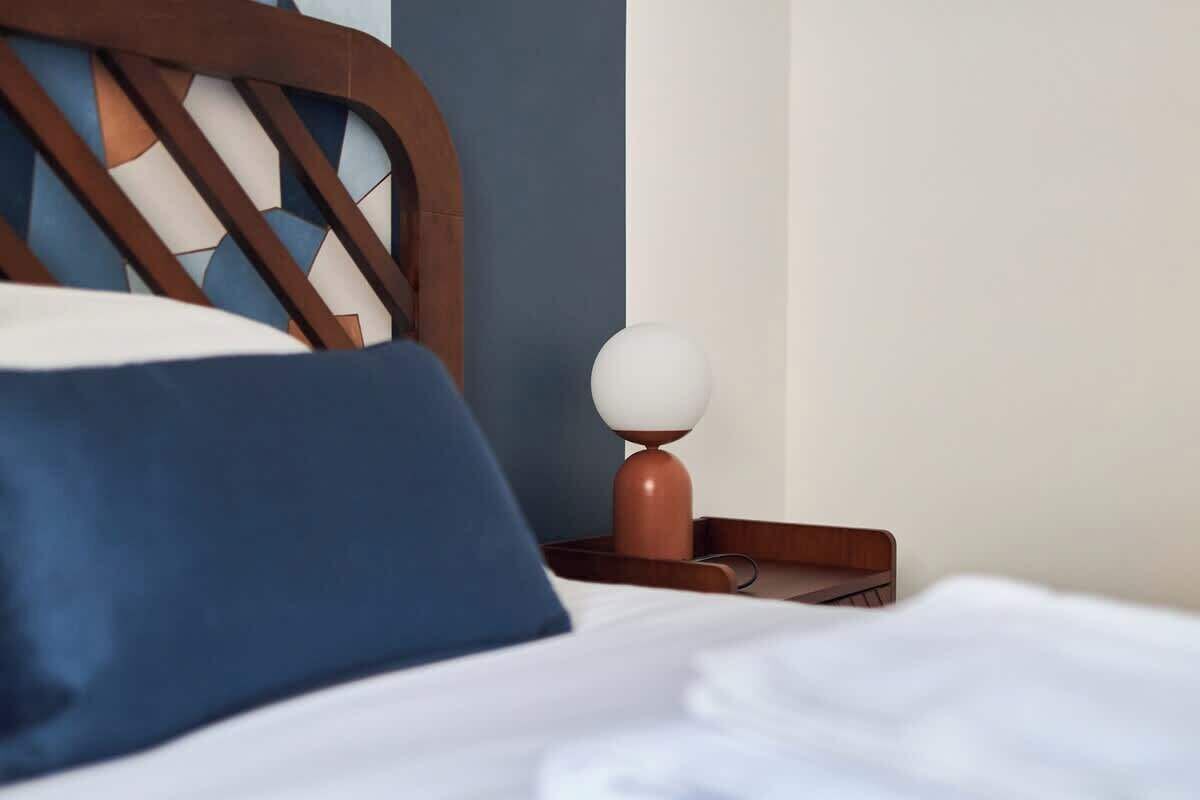 Un lit bien fait, avec une tête de lit en bois, un oreiller blanc et une table de chevet marron sur laquelle se trouve une lampe.