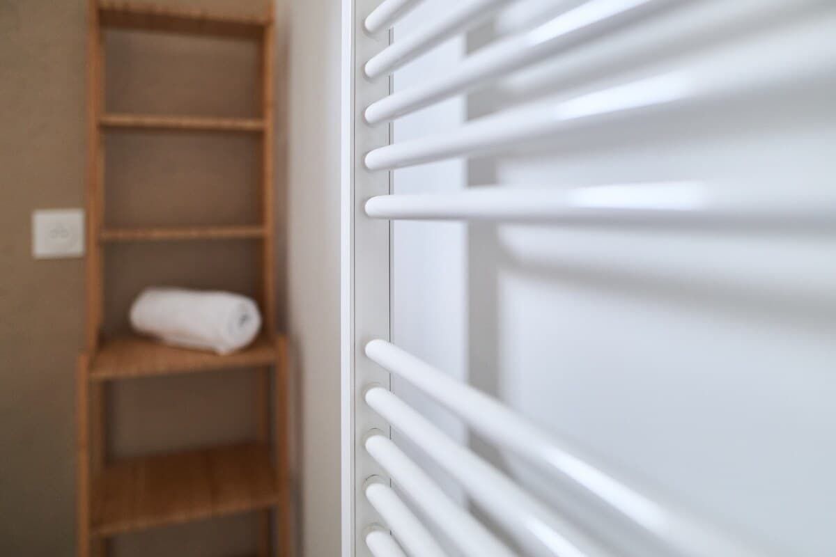 A white radiator with horizontal bars, a wooden shelf with a rolled towel, and a wall socket.