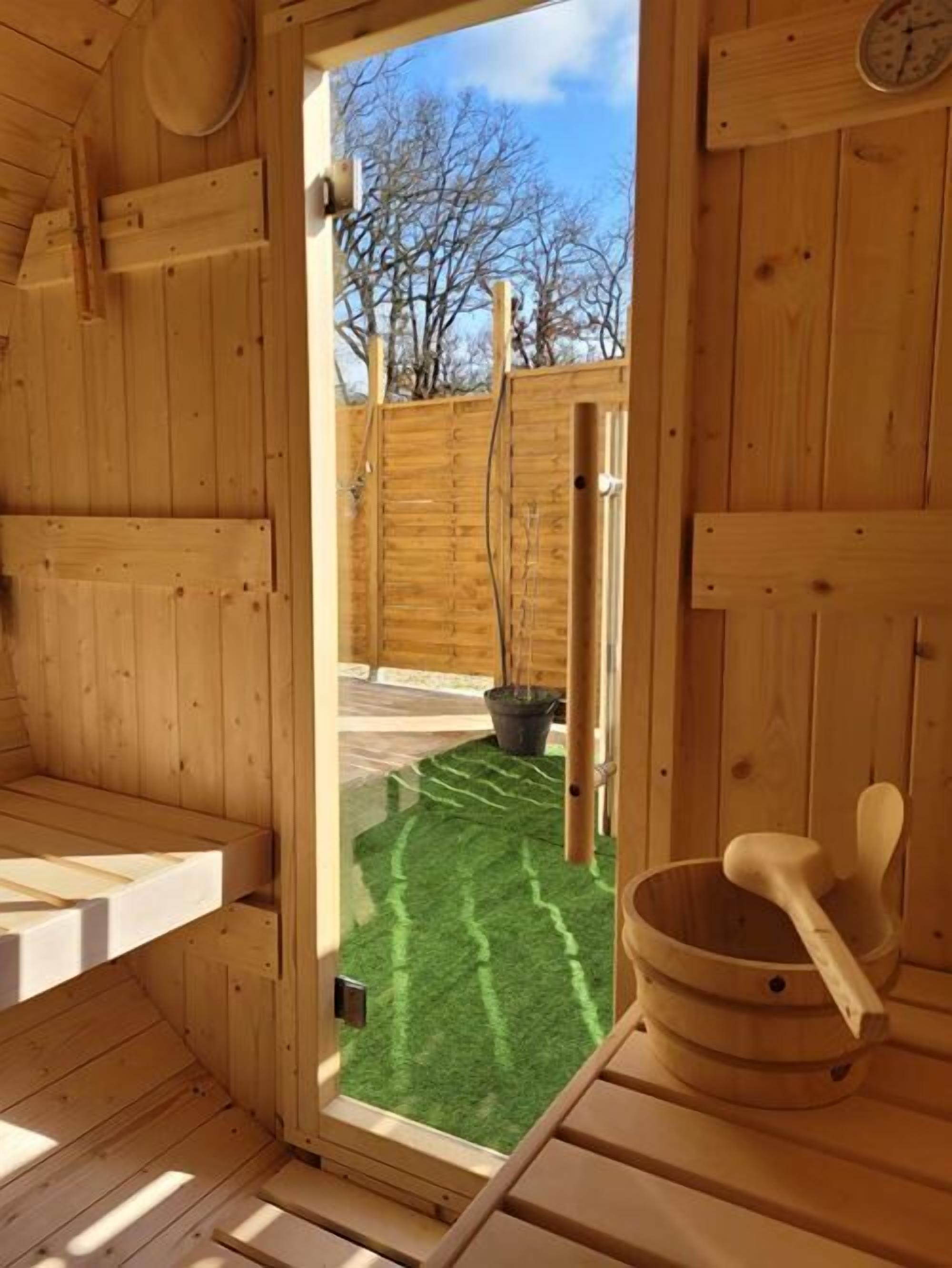 A wooden sauna room with a door leading outside, a wooden bench, a wooden bucket, and a small clock on the wall.