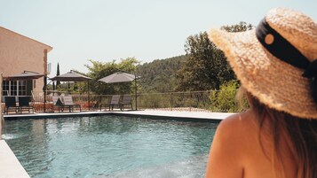 Zona junto a la piscina con sillones de descanso y sombrillas, una piscina cristalina y vistas a una ladera.