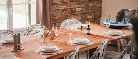 A wooden dining table set with white plates, glasses, and a bottle, surrounded by wicker chairs.