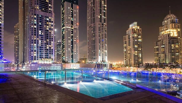 A nighttime view of a pool area with multiple swimming pools, surrounded by high-rise buildings with illuminated windows.
