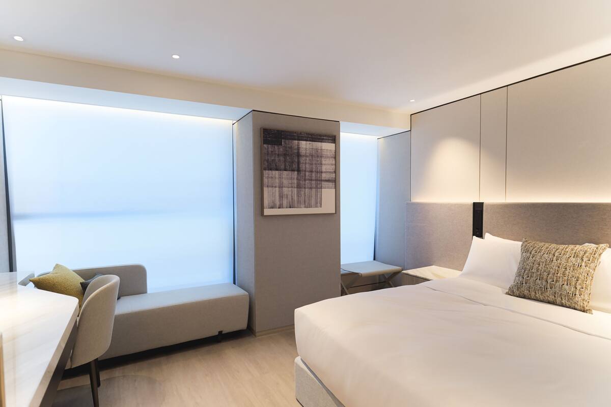 A modern hotel room with a large bed, a seating area with a chair and a small table, a large window, and a wall-mounted artwork.