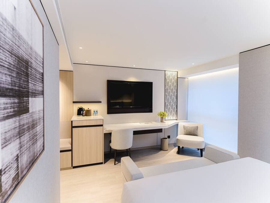 A modern hotel room with a minimalist design, featuring a large bed, a desk with a chair, a flat-screen TV, and a small seating area.