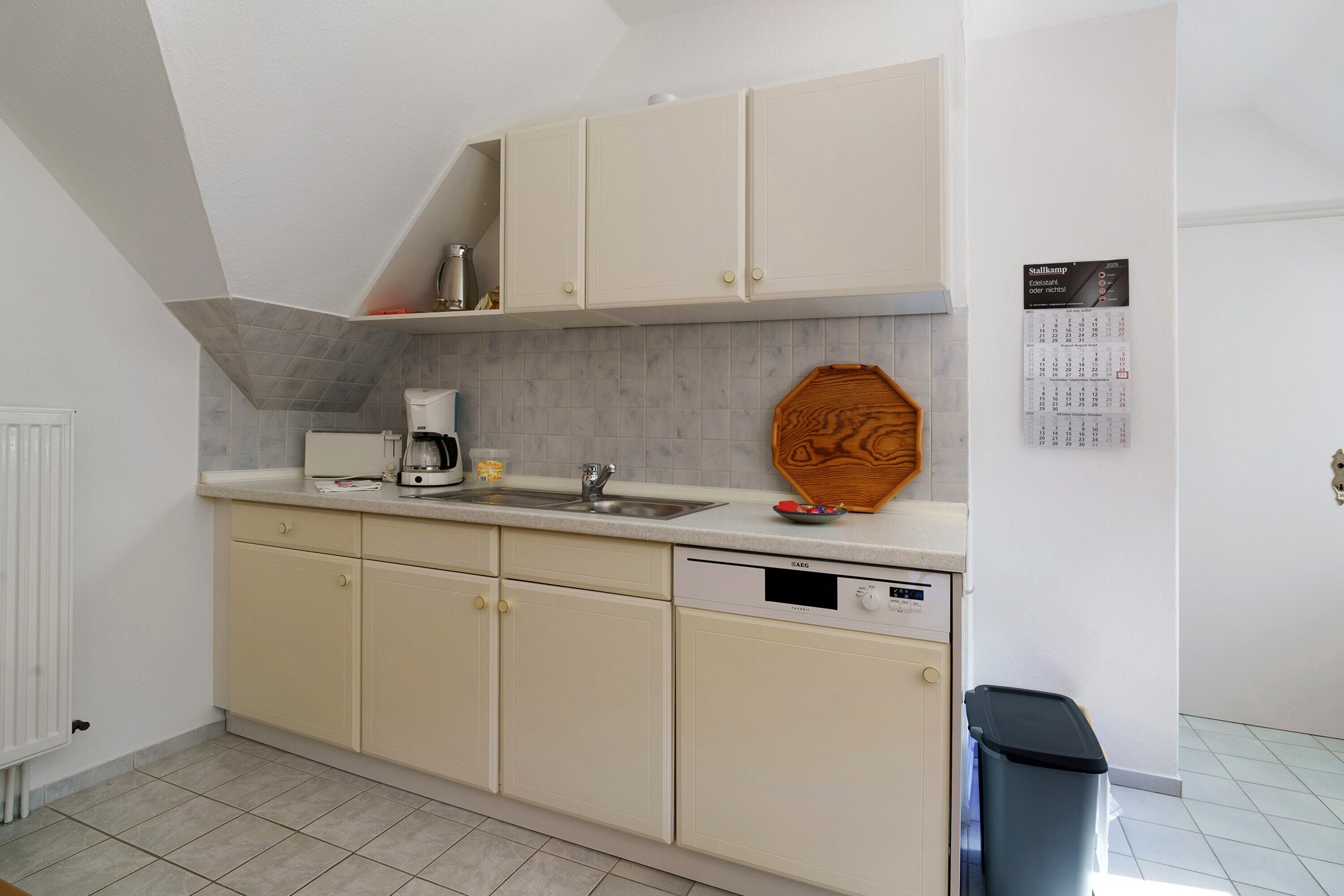A kitchen with white cabinets, a dishwasher, a coffee maker, and a calendar on the wall.