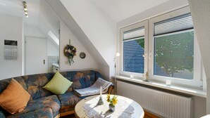 Ein Zimmer mit schräger Decke, einer Couch mit Kissen, einem runden Tisch mit Spitzenunterlage und einem Fenster mit Jalousien.
