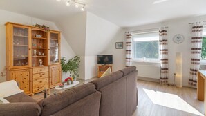 Ein Wohnzimmer mit braunem Sofa, Holzschrank, Fernseher und einem Fenster mit Vorhängen.