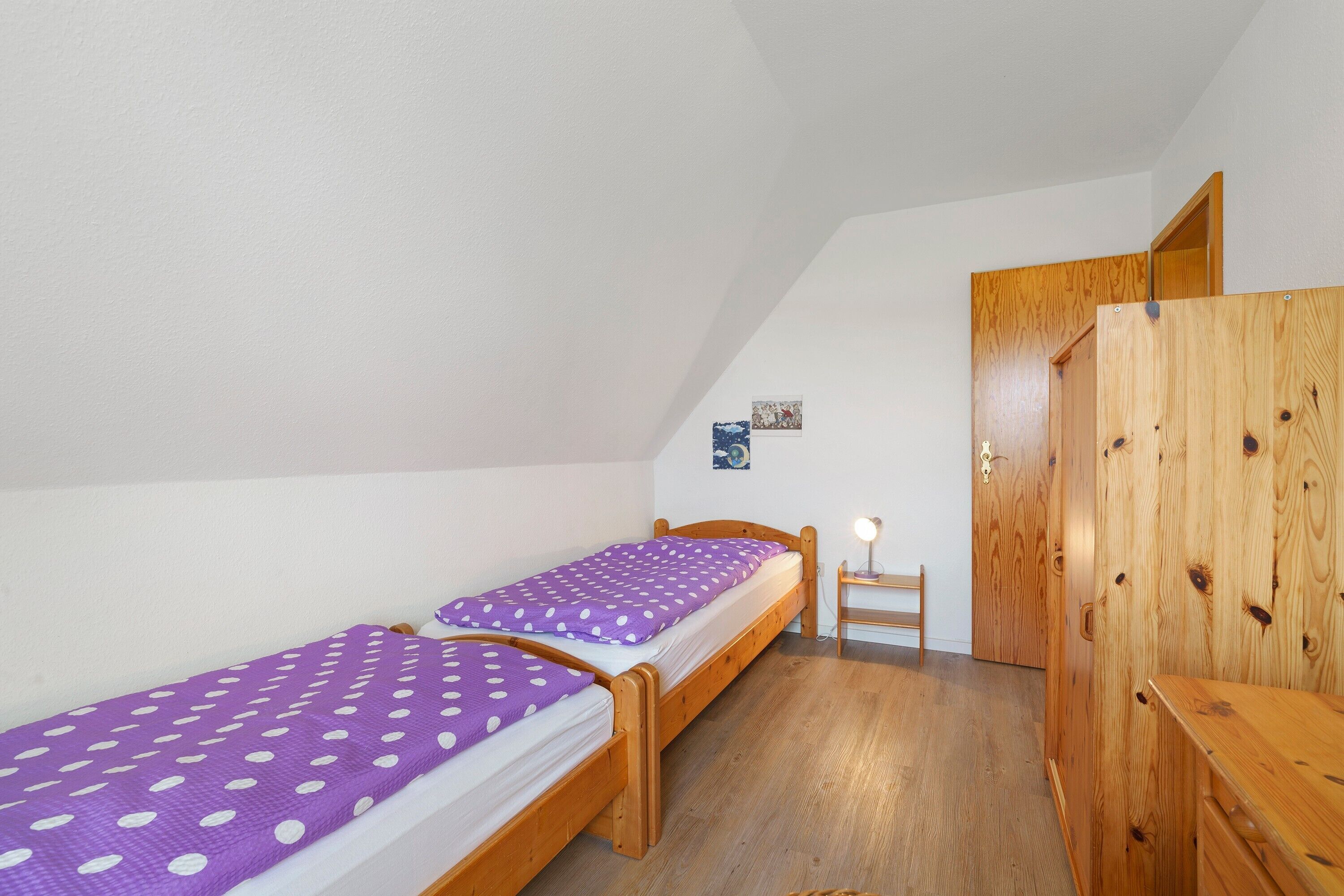 A small bedroom with two wooden beds, a wooden wardrobe, and a small table with a lamp.