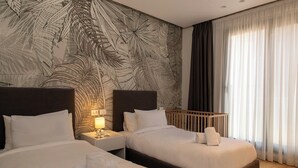 Habitación de hotel con dos camas, una mesita de noche con lámpara, una cuna y un amplio ventanal con cortinas translúcidas.