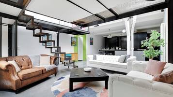 Un salon moderne avec un canapé en cuir marron, un canapé blanc et une table basse noire. Un escalier avec une rampe en métal mène à un autre niveau.