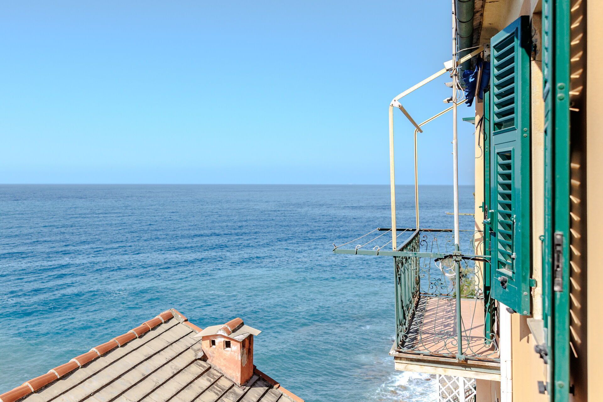 A balcony with a view of the sea, a tiled roof, and a building with green shutters.