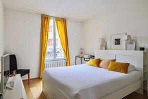 Une chambre d’hôtel avec un lit, un bureau, une chaise, une fenêtre avec des rideaux jaunes et un radiateur.