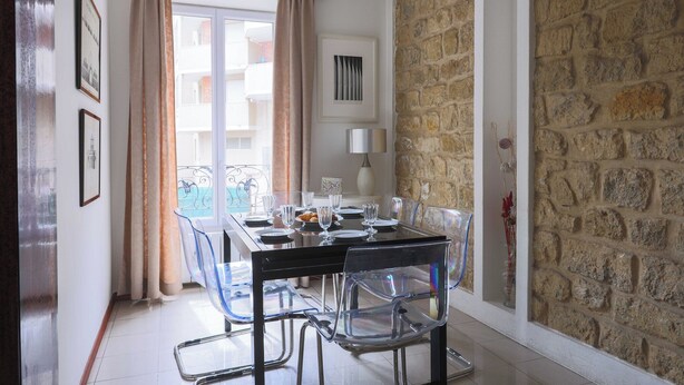 A dining area with a table set for four, clear chairs, a stone wall, and a view through a window with curtains.