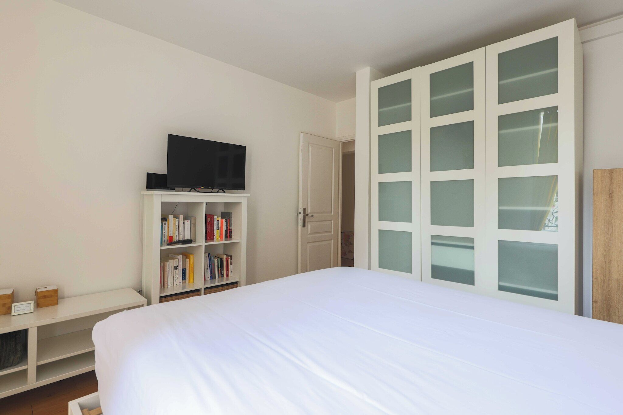 A bedroom with a bed, a television on a shelf, a bookshelf, and a wardrobe with glass doors.