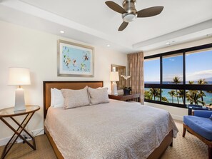 A hotel room with a bed, a ceiling fan, a large window with a view of the ocean, a painting on the wall, and a nightstand with a lamp.