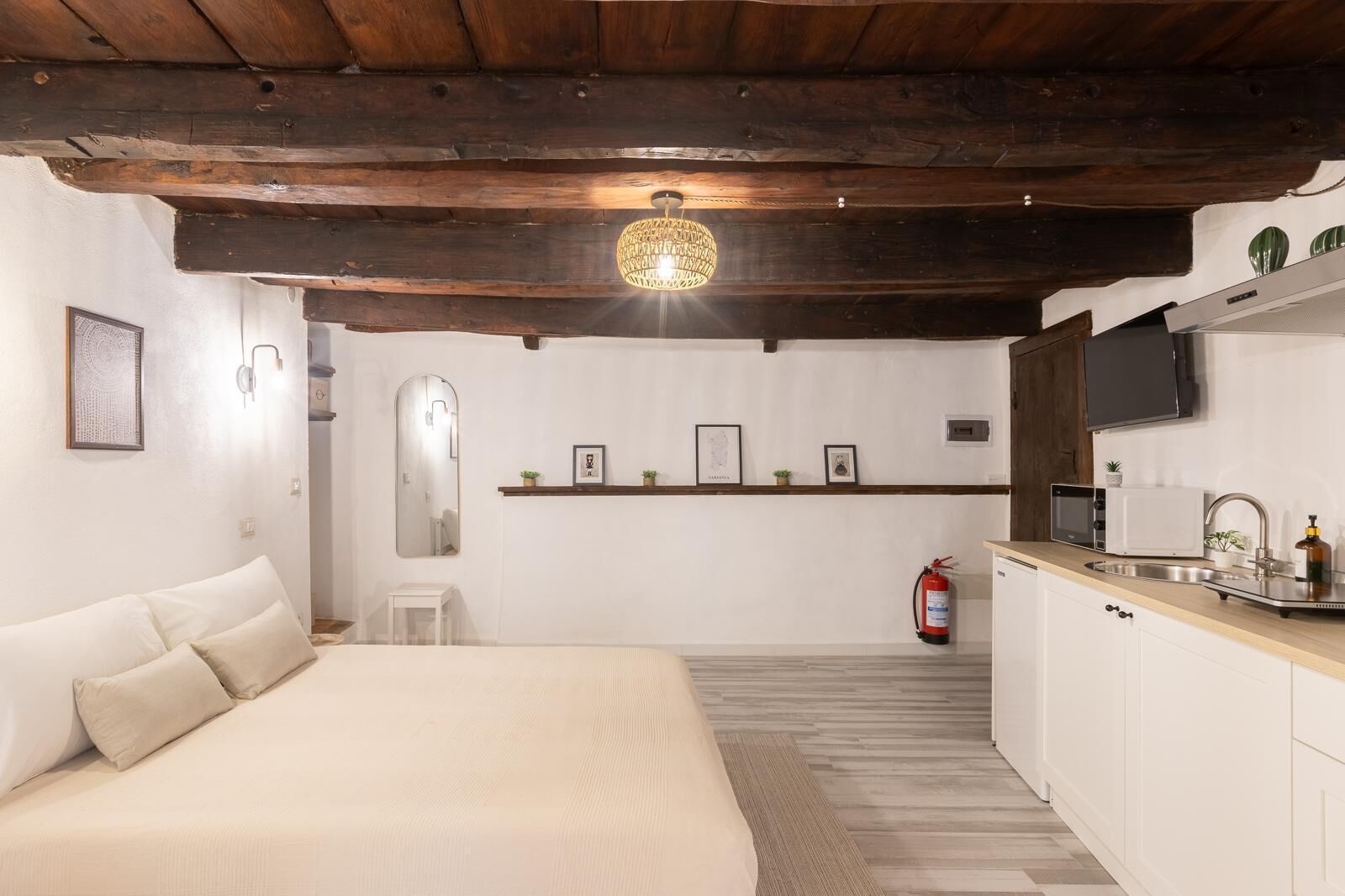 Un dormitorio con una cama, un taburete pequeño blanco, una mesita de noche, un espejo y una estantería con marcos de fotos.