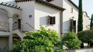 Un immeuble de deux étages avec une façade blanche, des volets en bois et un toit recouvert de tuiles. On y trouve des balcons avec des garde-corps et un escalier menant à l’étage supérieur.