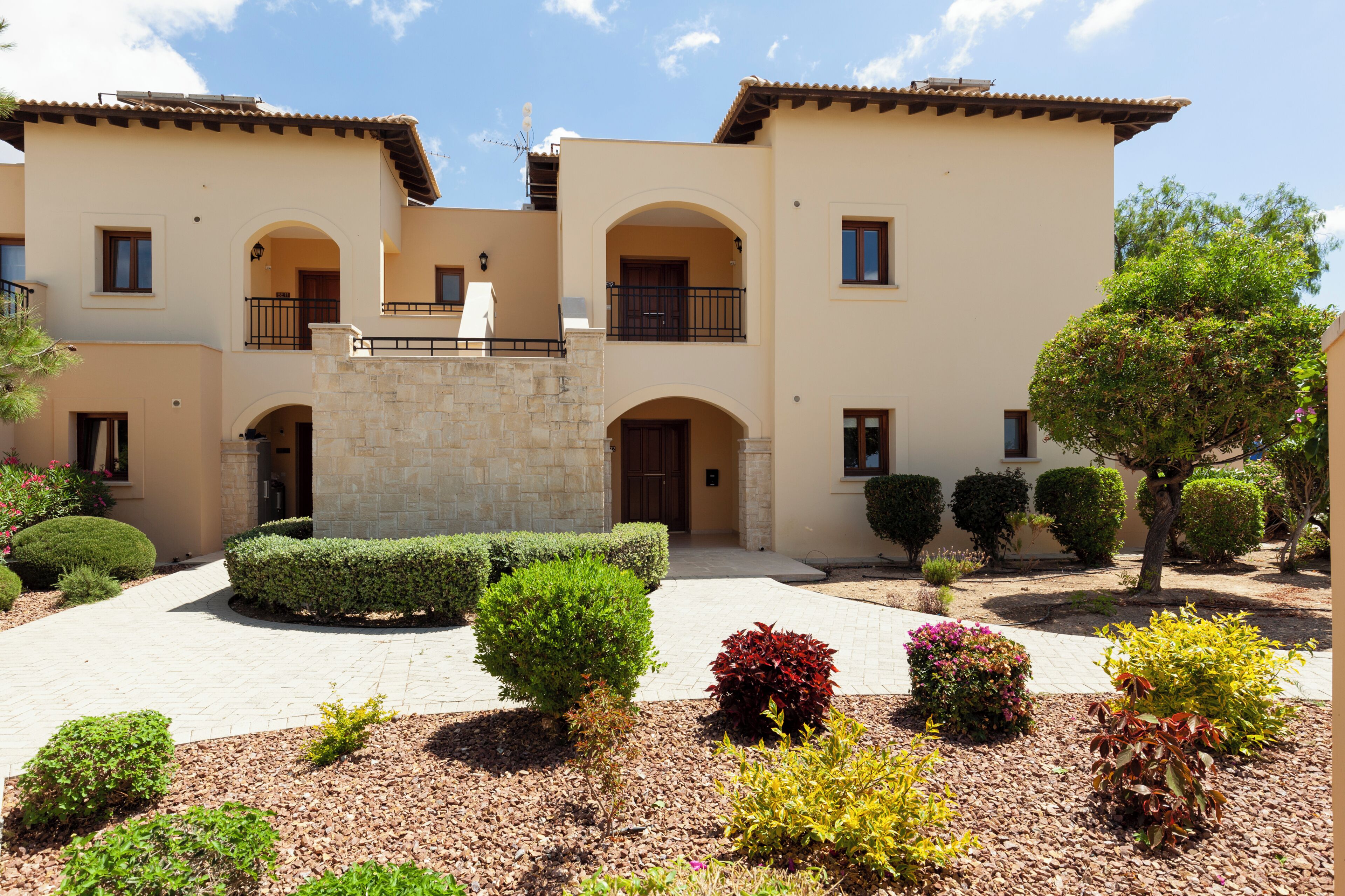 A two-story building with a stone facade, multiple balconies, and a landscaped courtyard with various shrubs and plants.