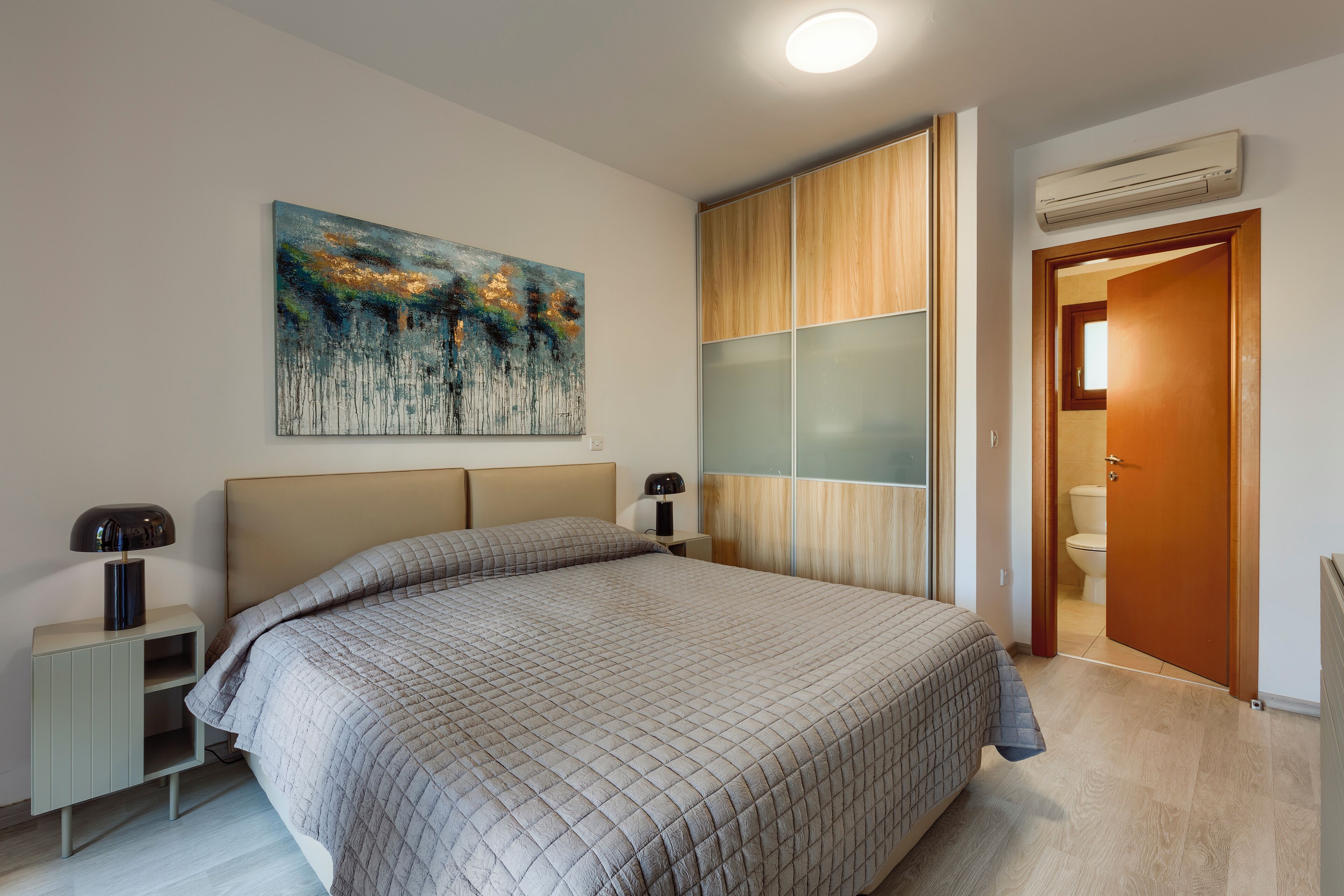 A bedroom with a large bed, a nightstand with a lamp, a wall-mounted air conditioner, and a door leading to a bathroom.